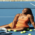 Lady smiling and sitting on a tennis court wearing an orange dress next to tennis balls.