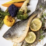 A picture of a fish dish with lemon slices and herbs plated next to roasted vegetables.