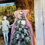 A Christmas tree window display next to a clothed mannequin.