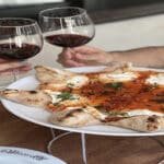A couple toasting glasses of red wine next to a fancy pizza with fresh mozzarella.