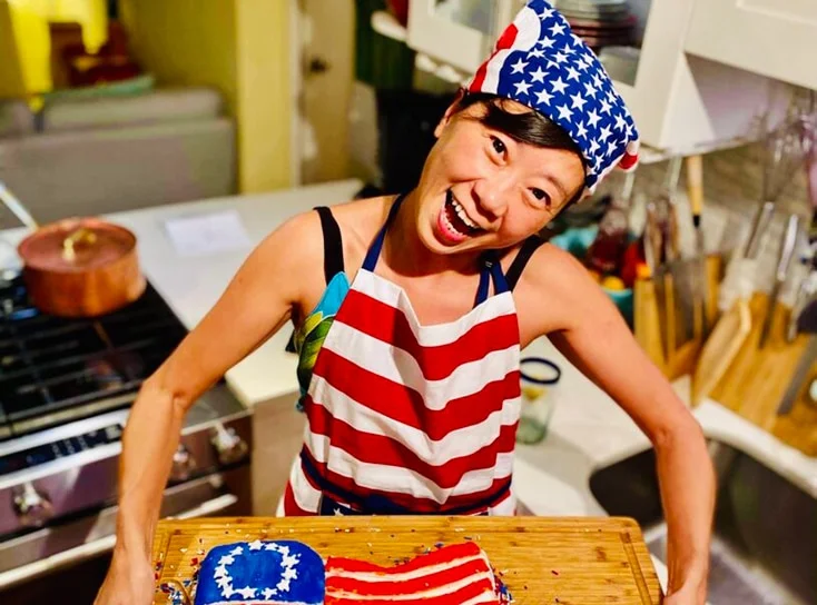 A lady wearing an American flag bandana and apron, smiling holding a board with an American theme cake.