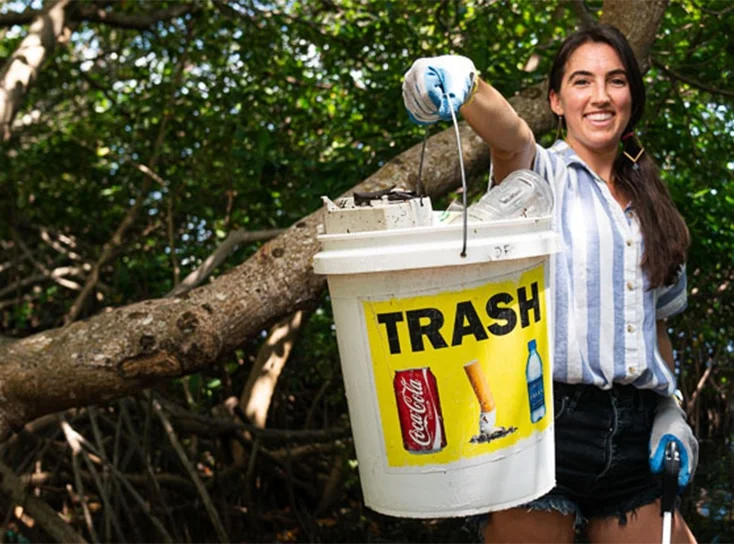 A young lady smiling and holding a white bucket with a yellow label that says trash. She is wearing gloves and a blue and white stripped blouse.