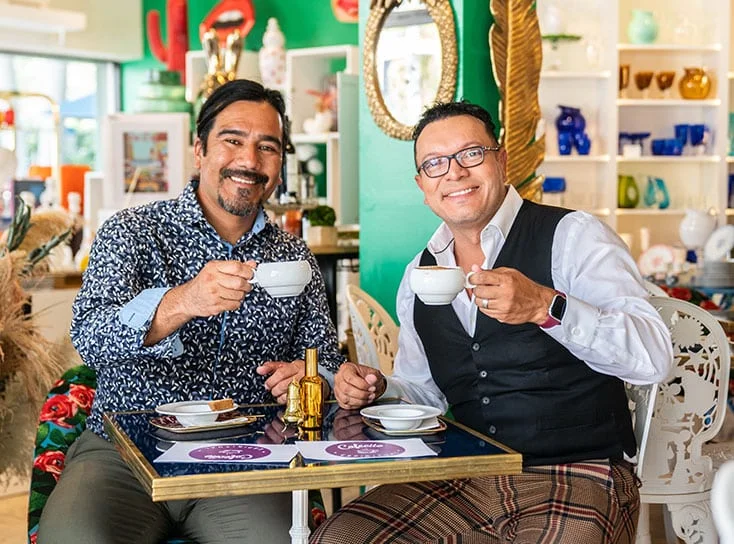 Two men sitting a table smiling holding a cup of coffee.