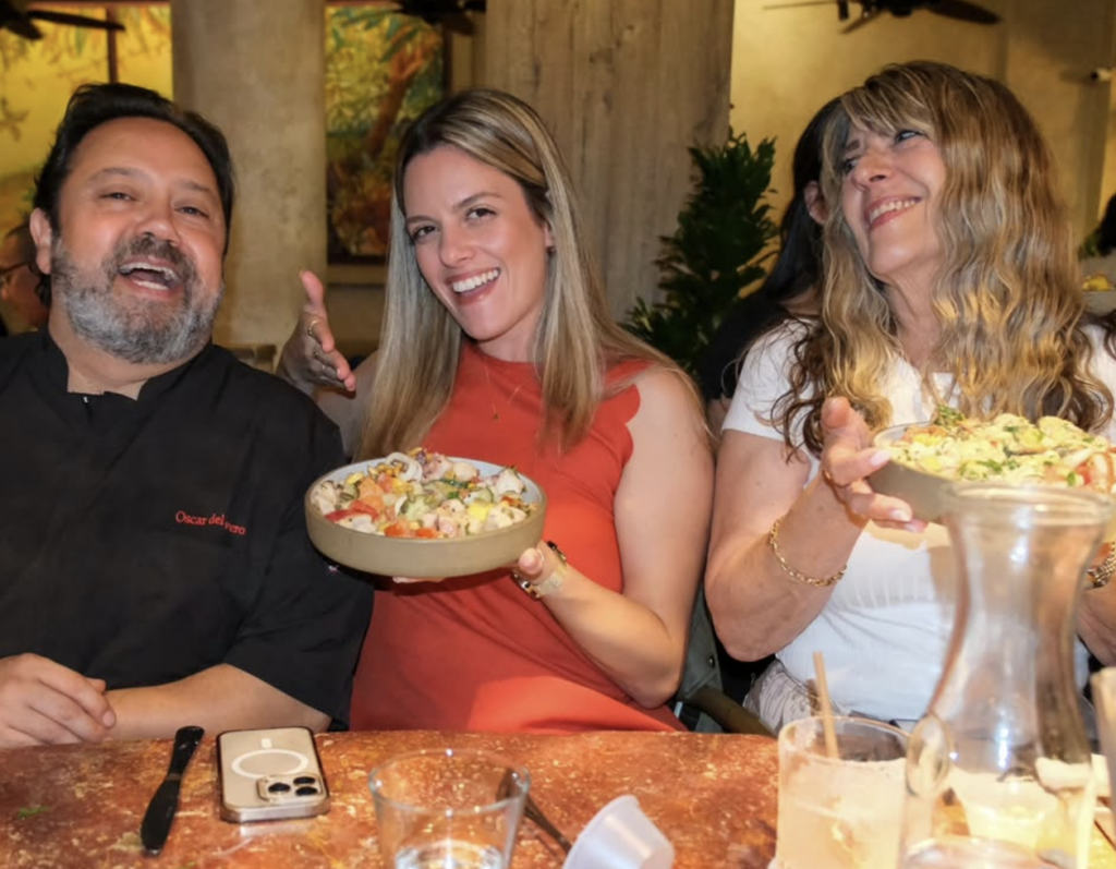 two happy women holding plates of food, next to a chef sitting at a restaurant.