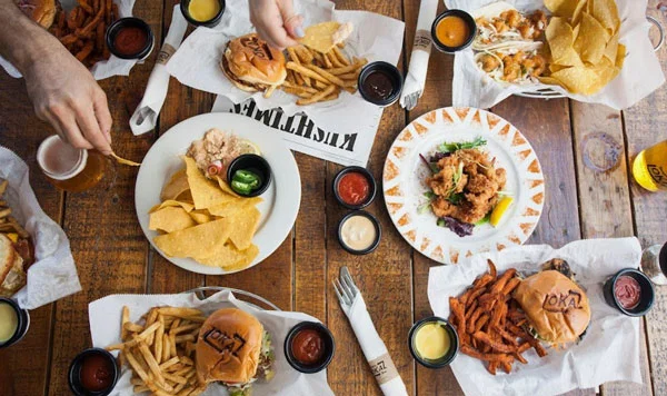 Multiple bar food dishes of burgers with fries and condiments on a wooden table.