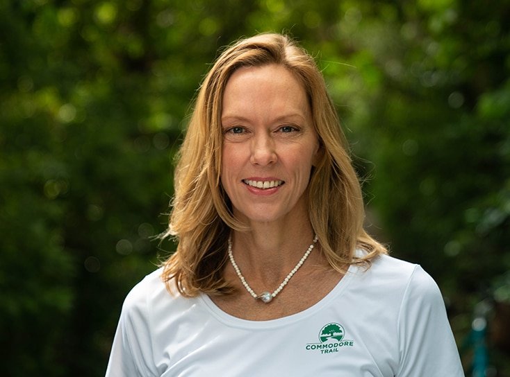 A lady smiling in a photograph outside wearing a white shirt with the commodore trail logo.