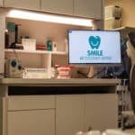 Dentist office with computer and patient chair next to a tv displaying SMILE Dentisty logo.
