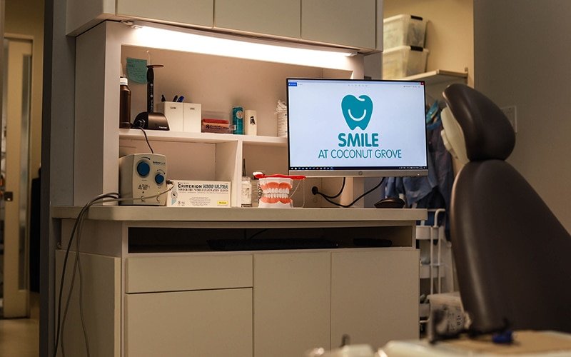 Dentist office with computer and patient chair next to a tv displaying SMILE Dentisty logo.