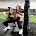 A lady assisting another lady in a squatting exercise with a barbell and weights inside of a gym.