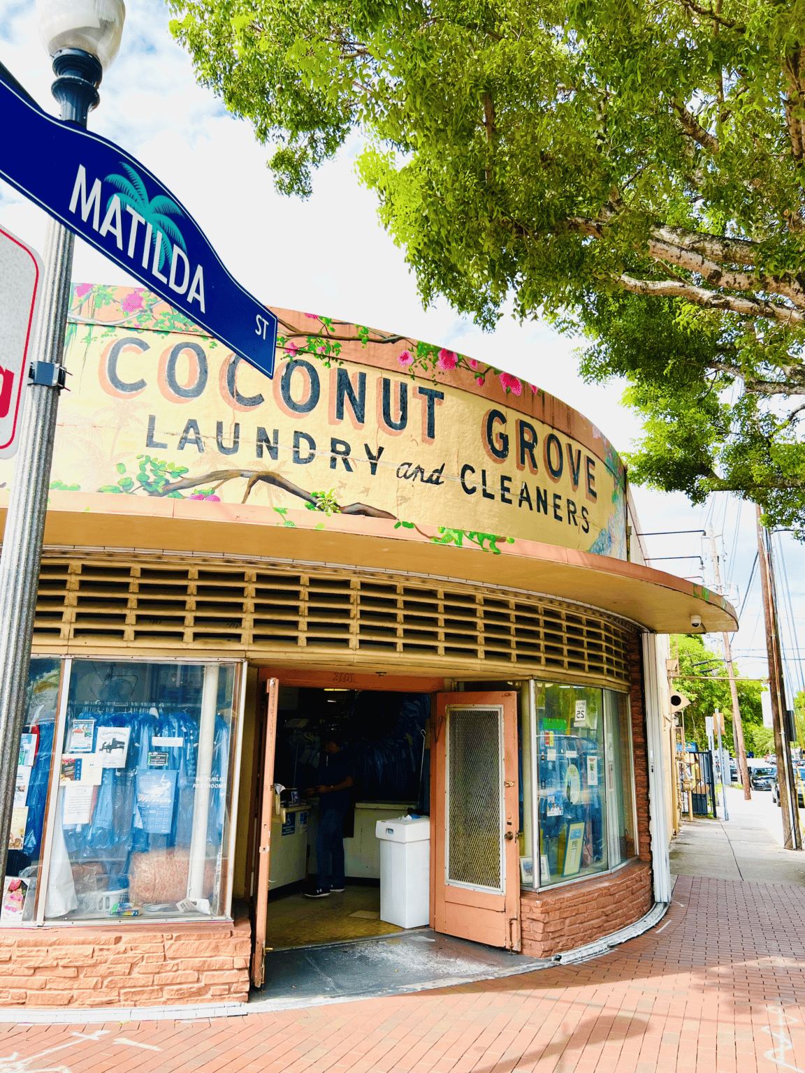 The exterior building of Coconut Grove Cleaner’s next to a tree and street sign.