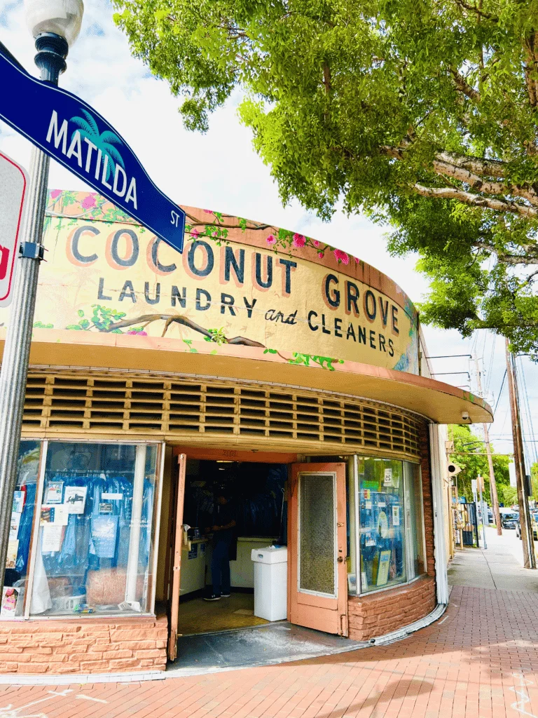 The exterior building of Coconut Grove Cleaner’s next to a tree and street sign.