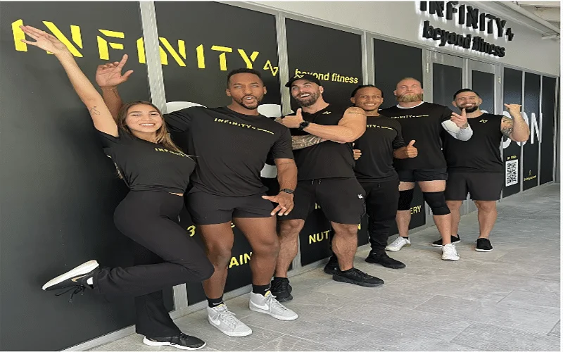 Group of people standing in front of a wall sign, wearing black workout uniforms, for Infinity Fitness.