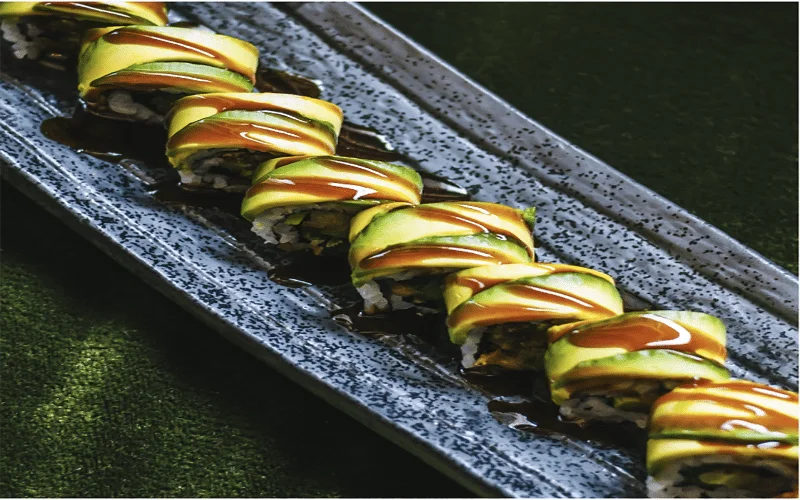 Sushi dish topped with avocado and plated a fancy long dish from PLANTA Queen, Miami. Opened to create delicious food that promotes environmental sustainability.