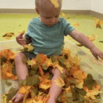 A little boy wearing a blue shirt is playing with leaves inside of Silly Monkeys Playhouse.
