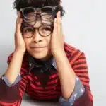 A boy with two pairs of glasses on his face. He is wearing a red stripped shirt.