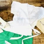 A white blouse and sandals with a green pair of shorts, laid out flat on a wooden table next to a tote bag that states 