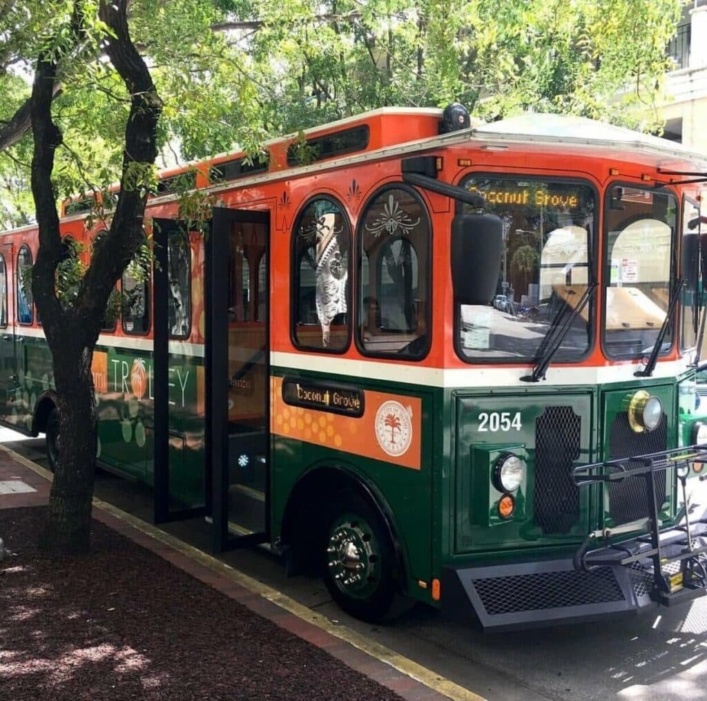 Trolley car in Coconut Grove, Florida, stopped next to a sidewalk and trees.