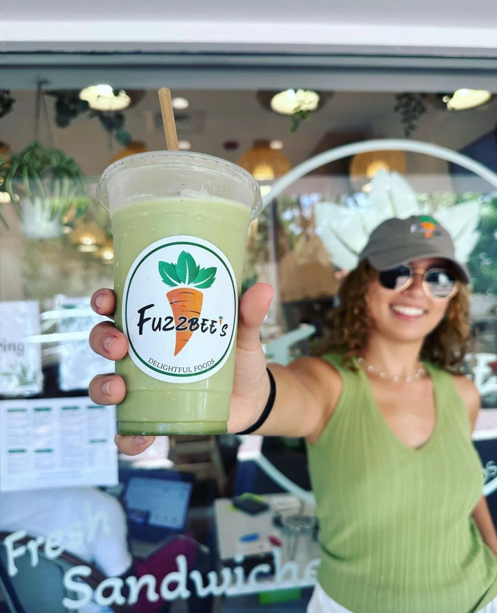 girl in green basebal cap and green tank top holds a fruit smoothee in a cup with a Fuzzbee's carrot logo