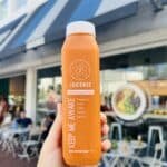An orange colored smoothie in a bottle being held up by a person in front of a smoothie bar, Juicense.