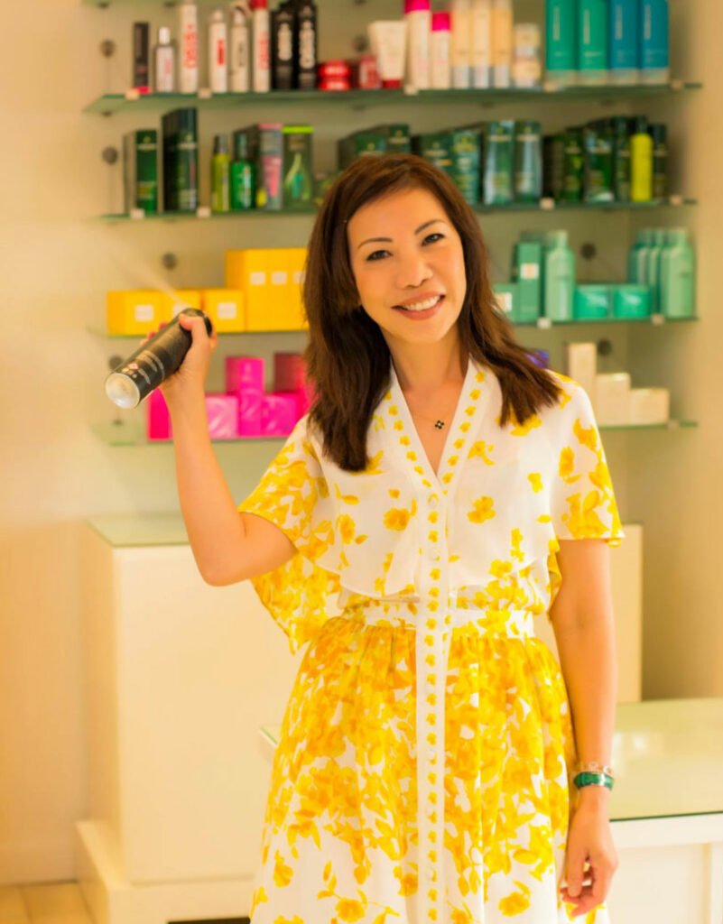 A lady wearing a white and yellow floral dress, holding a bottle of shampoo and smiling.