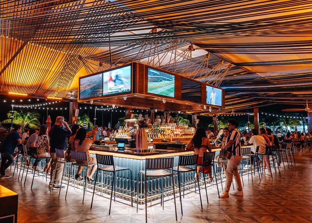 A bar with large TVs above it and the bar is surrounded by people socializing.