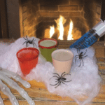 Halloween themed drinks sitting on a table with fake spider webs and next to a fireplace.