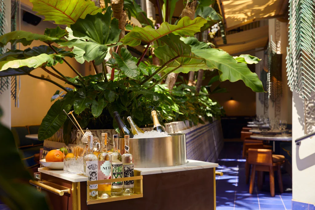 A rolling bar cart with drinks and garnishes next to potted plants in the Fountain Lounge.