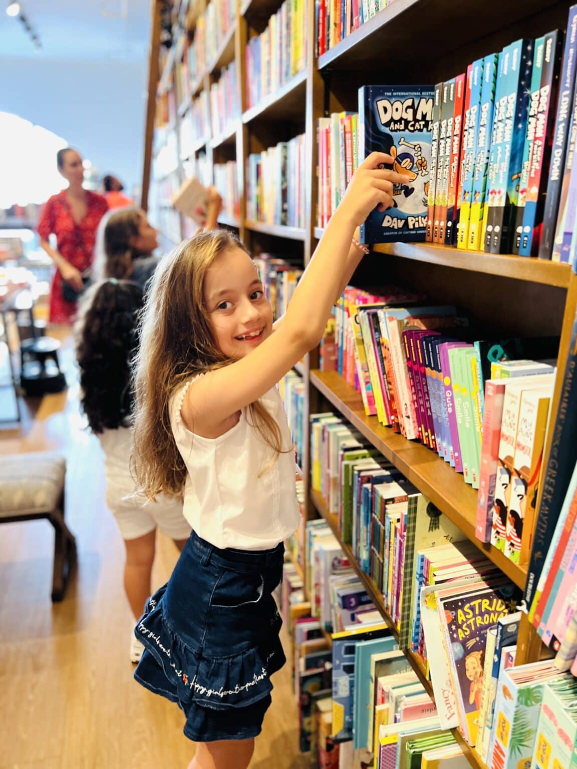 A young girl browsing through a shelf of books, reaching for a book.