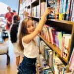 A young girl browsing through a shelf of books, reaching for a book.