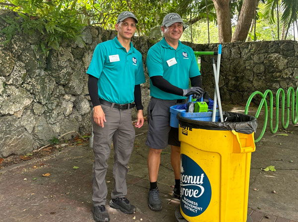 two men in teal polo shirts with rakes and brooms and yellow garbage receptacle