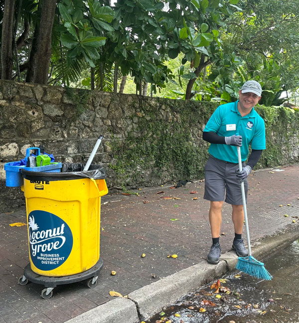 Worker cleaning street in Coconut Grove with a broom and garbage can.
