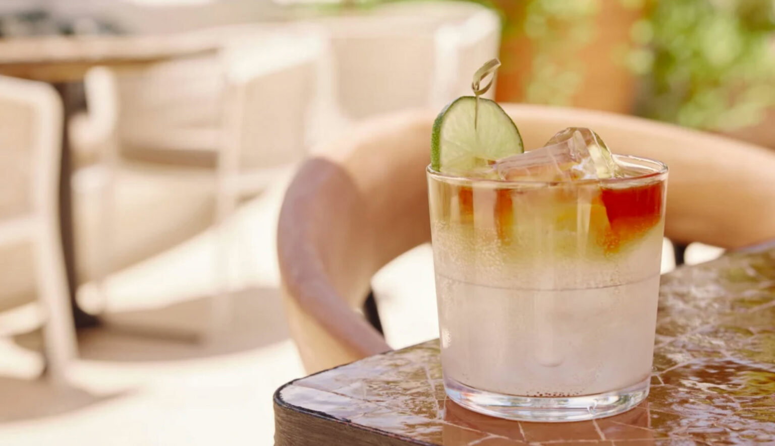 A lime garnished brown and white drink with ice sitting on a tiled table outside at a restaurant.