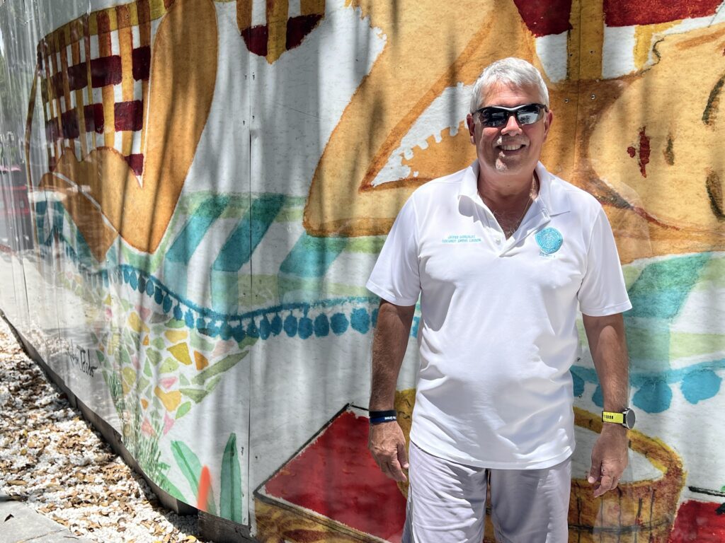 Man wearing sunglasses smiling next to a painted wall mural outside.