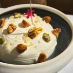 Honey being drizzled on whipped dessert topped with a variety of nuts and edible flowers.