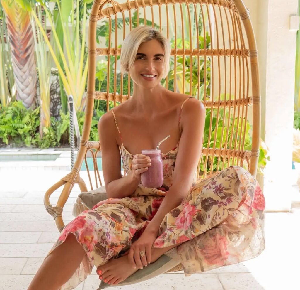 Lady sitting in a chair holding a smoothie in a glass jar. With tropical greenery behind her.