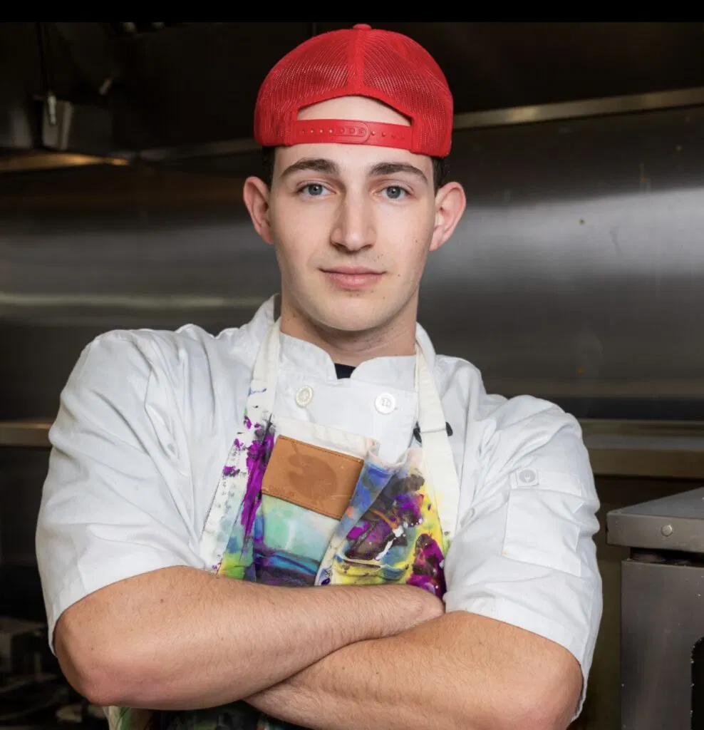 chef posing for a photo in uniform and colorful apron with a backwards red hat.