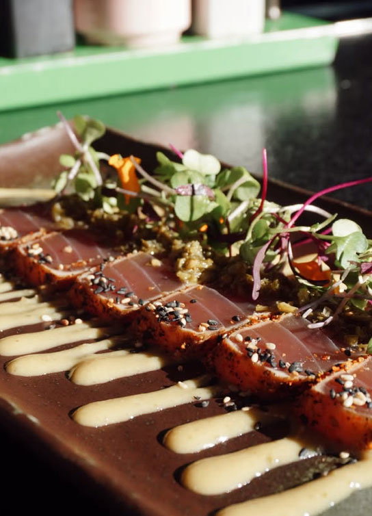 a plate of raw fish from Sushi garage topped with micro greens, and sesame seeds, and white sauce.
