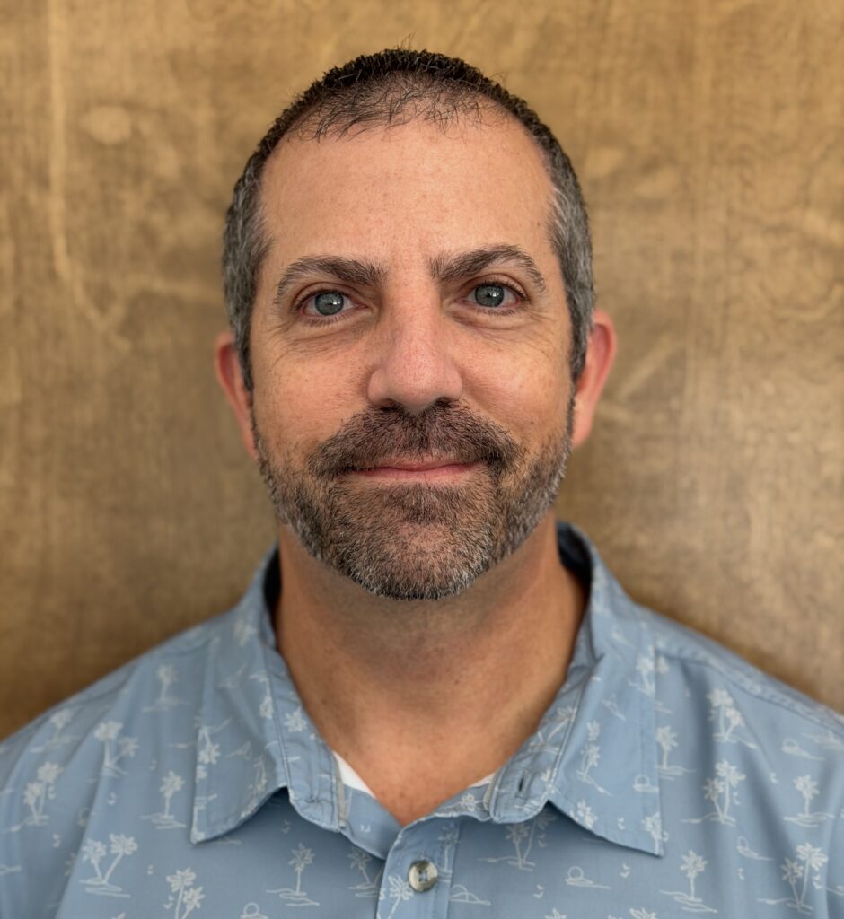 man smiling for headshot photo wearing a palm tree button up.