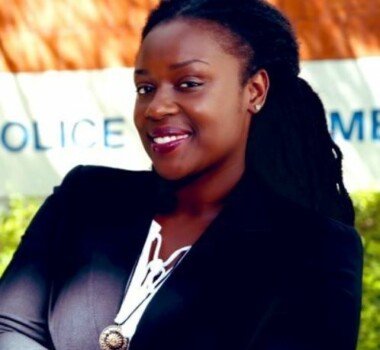 A young lady smiling in front of the police building while wearing a black suit.