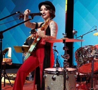 A lady wearing red outfit singing in a microphone and playing a guitar.